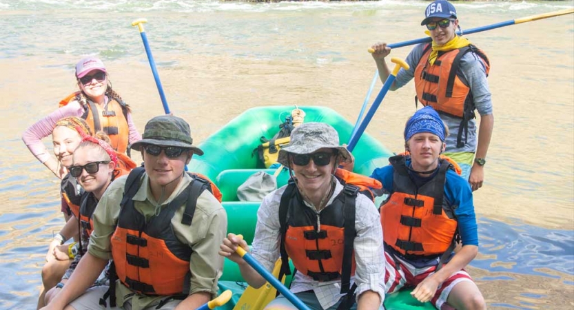 A group of people wearing life jackets sit in or stand near a raft floating in shallow water and smile at the camera